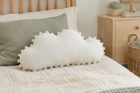 Coussin décoratif blanc en coton, en forme de nuage avec des pompons, posé sur un lit d’enfant