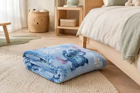 Plaid polaire bleu clair à motif Stitch posé au pied d’un lit d’enfant dans une chambre lumineuse.